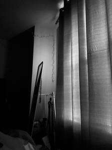a black and white photo of the inside of a bedroom taken from the bed, the curtains are drawn light only partially pouring through revealing the backside of a standing mirror, the room fading to black.