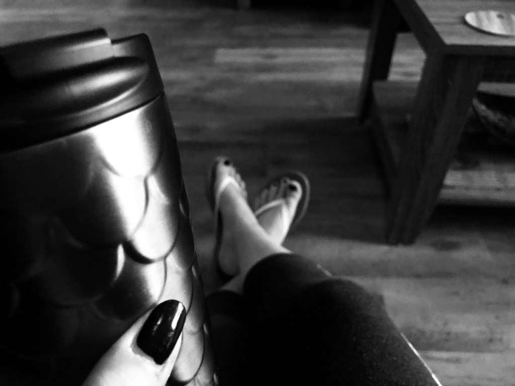 a scalloped coffee thermus being held by a hand with black nail polish, in the distance feet are stretched out into room