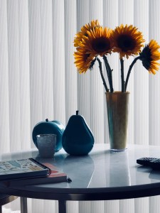a color photo of vibrant yellow sunflowers in a cup next to blue ceramic apples and pears sculptures 