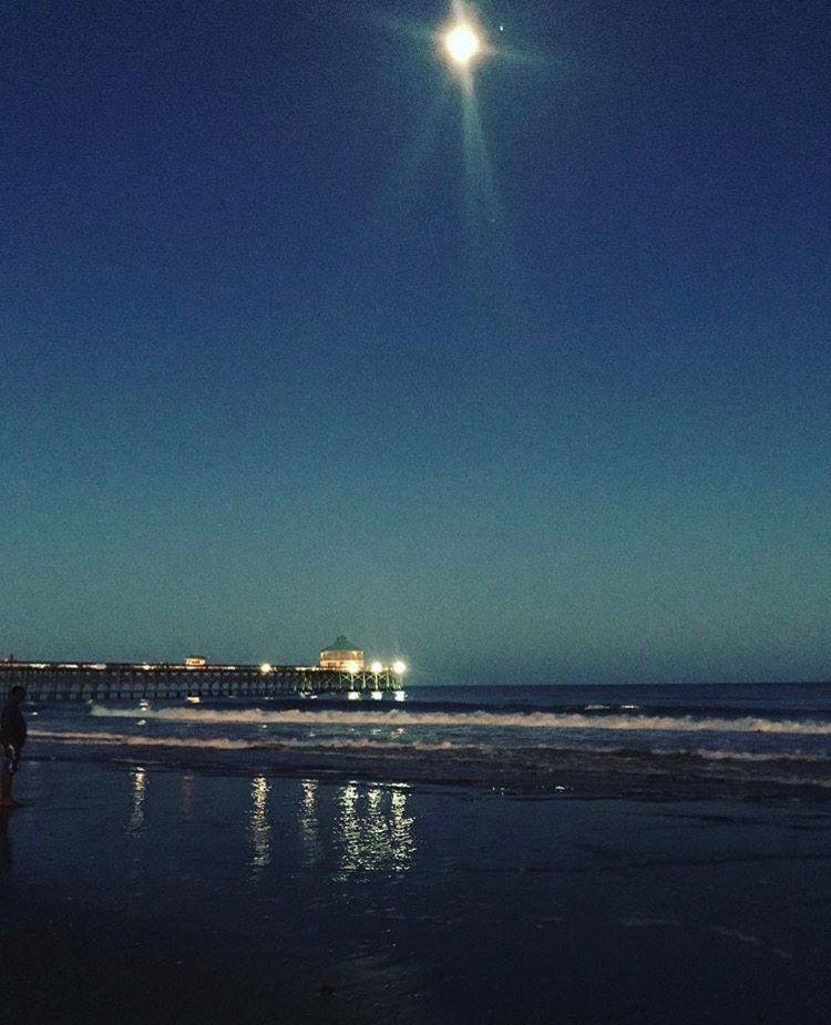A very clear blue night sky with the full moon shining bright over the top of an illuminated pier extending over the ocean.