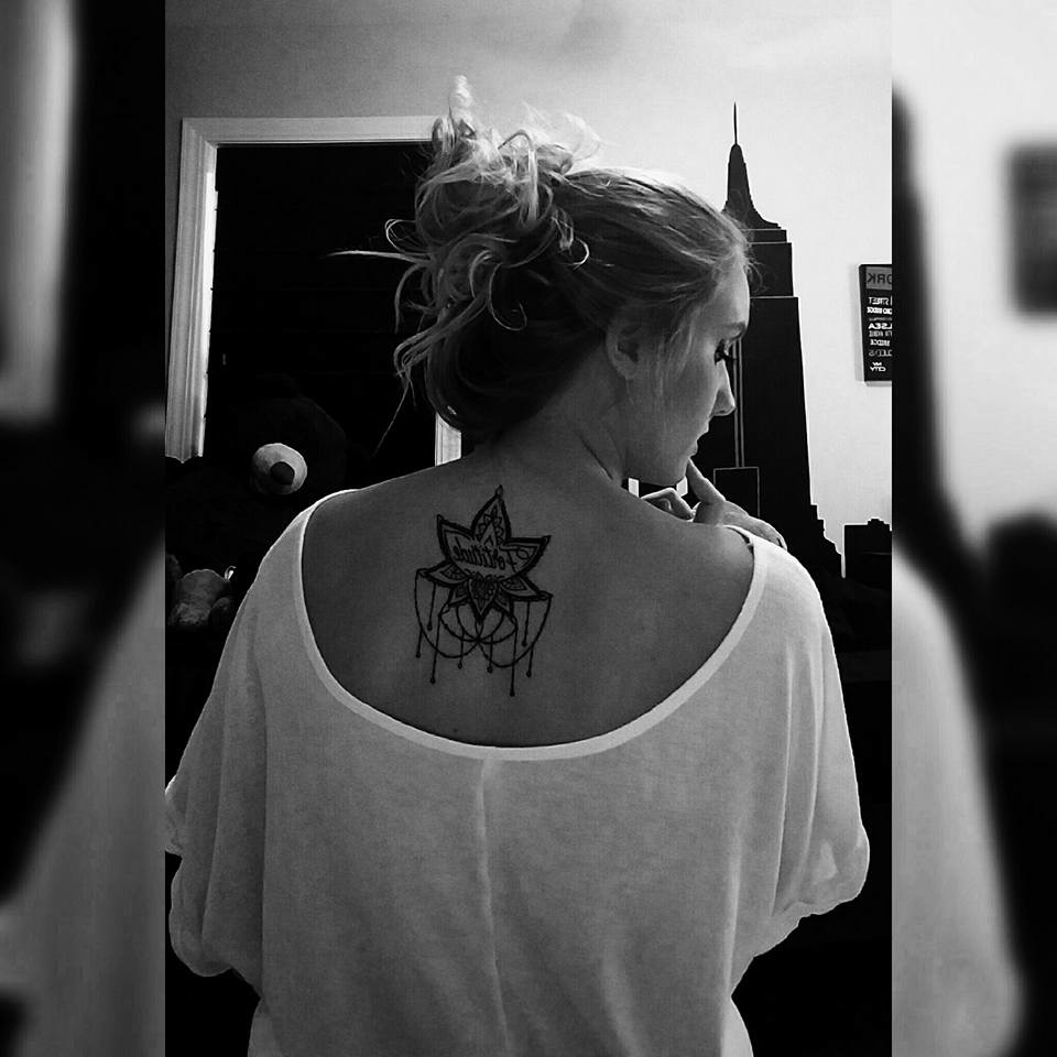 black and white photo of girl with hair pulled up, facing away from the camera. Her white blouse reveals a tattoo of a lotus flower on her back inscribed with "fortitude"
