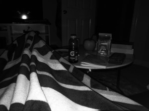 a black and white photo of my legs outstretched covered in a fuzzy packers blanket in a dark room with a gatorade on my table 