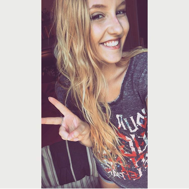 photo of white woman with long wavy blonde hair, sunkissed skin and visible freckles throwing up a peace sign while smiling and wearing a def leppard tshirt