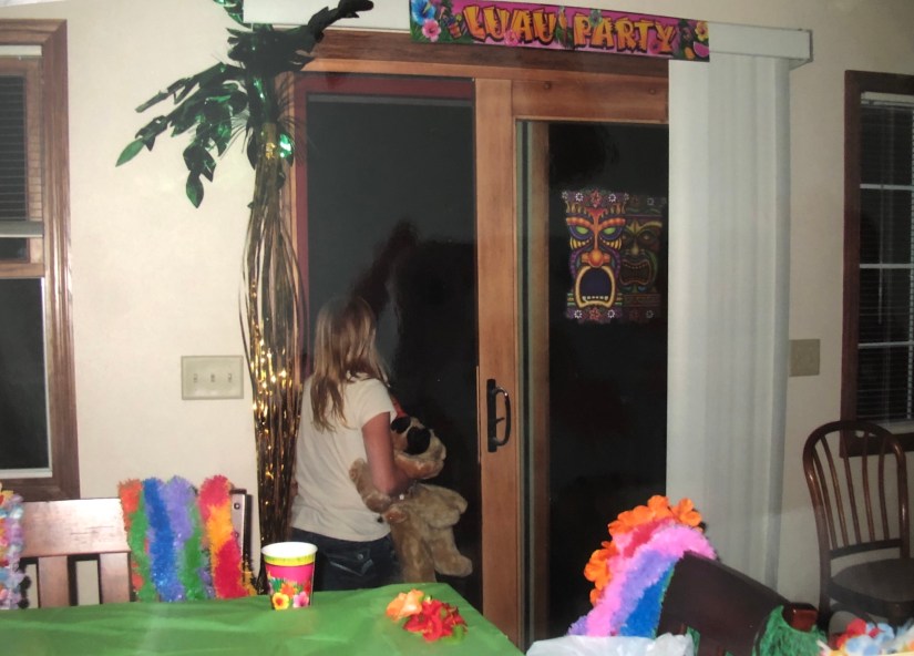 a photo of me as a kid, facing a dark patio door tightly holding my stuffed dog with a lua theme party behind me watching the storms