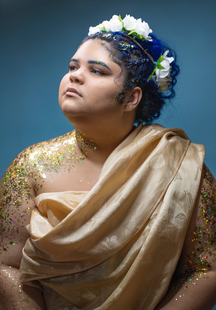 A photo of Jude Valentin, a body positivity advocate. She sits looking away from the direct camera, her blue hair pulled back by a flower crown. Her body adorned with gold glitter and draped with a gold cloth like a grecian goddess