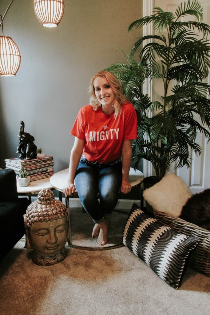 photo of blonde woman with medium blonde hair sitting on a marble coffee table surrounded by pillows, plants, records and spiritual items.