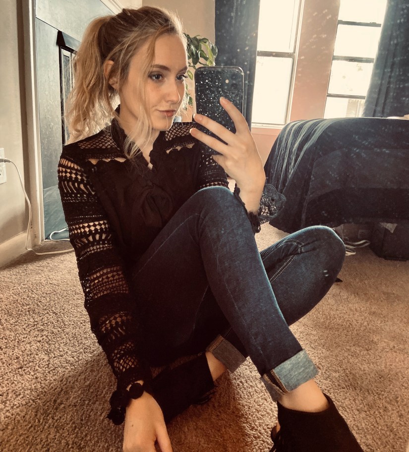 Girl with blonde hair tied back in a pony tail sitting on the floor wearing a nice black blouse, jeans, and black heeled booties. 