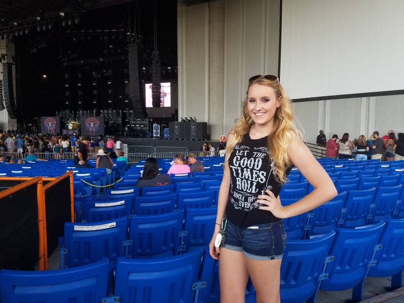 Blonde girl with rock n roll t-shirt on standing in front of a slowly filling concert venue. 