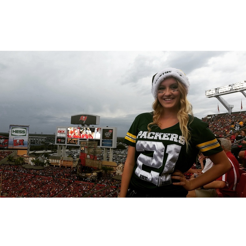 Blonde girl wearing a packers christmas hat and jersey with the Tampa Bay Buccaneers stadium in the background. 