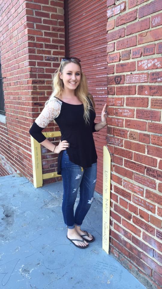 Girl in a black top and jeans standing against a brick wall on the set of One Tree Hill. 