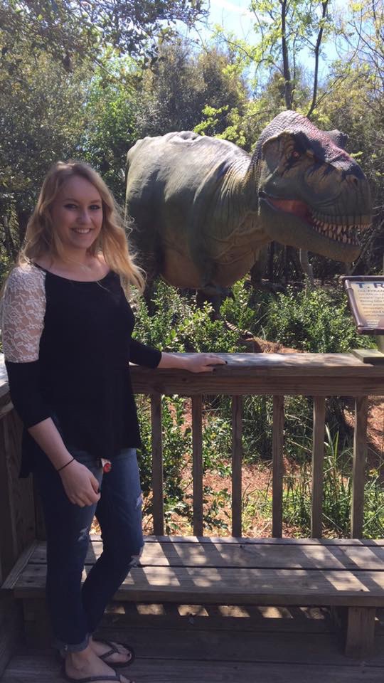 Blonde in a black shirt posing in front of a T-rex behind a fence. 