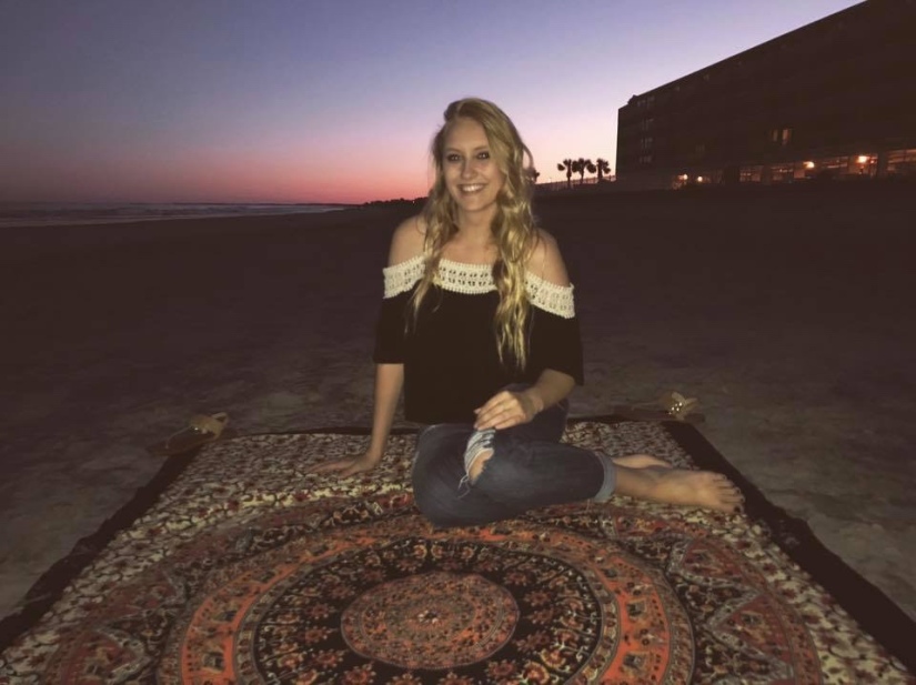 Blonde girl in summery black shirt with lace trim sitting on a mandala printed tapestry on the beach with the sun setting behind her. 