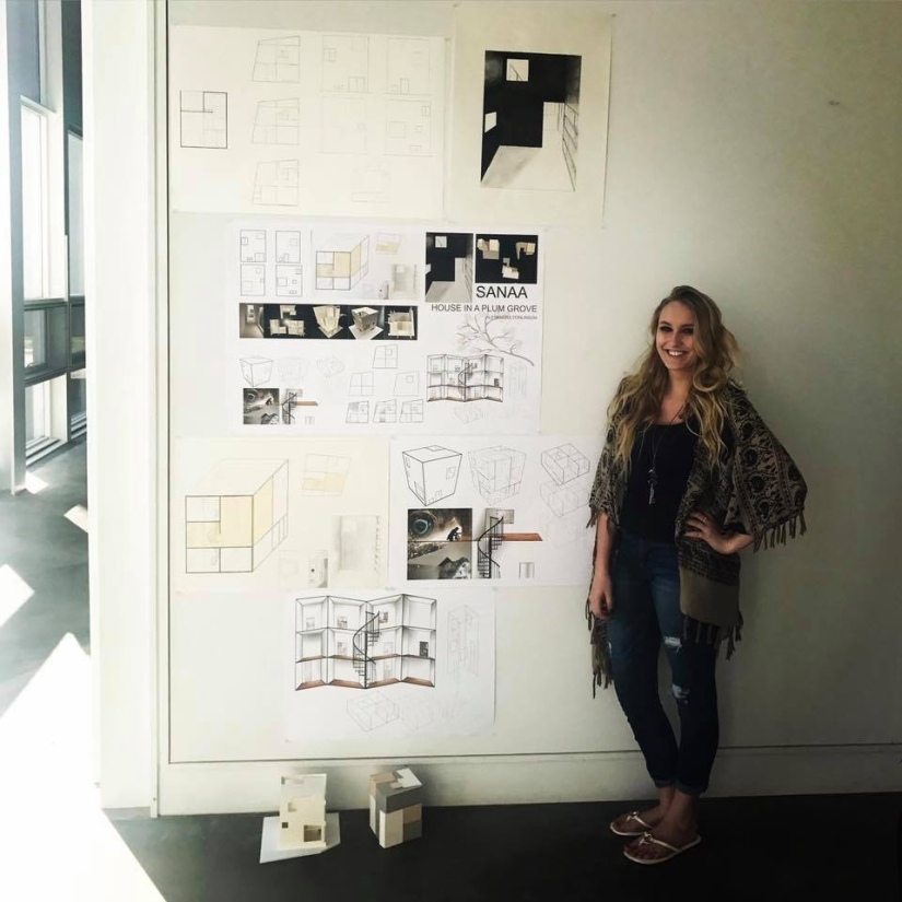 Girl standing next to a large wall full of architectural drawings on display with models on the floor.
