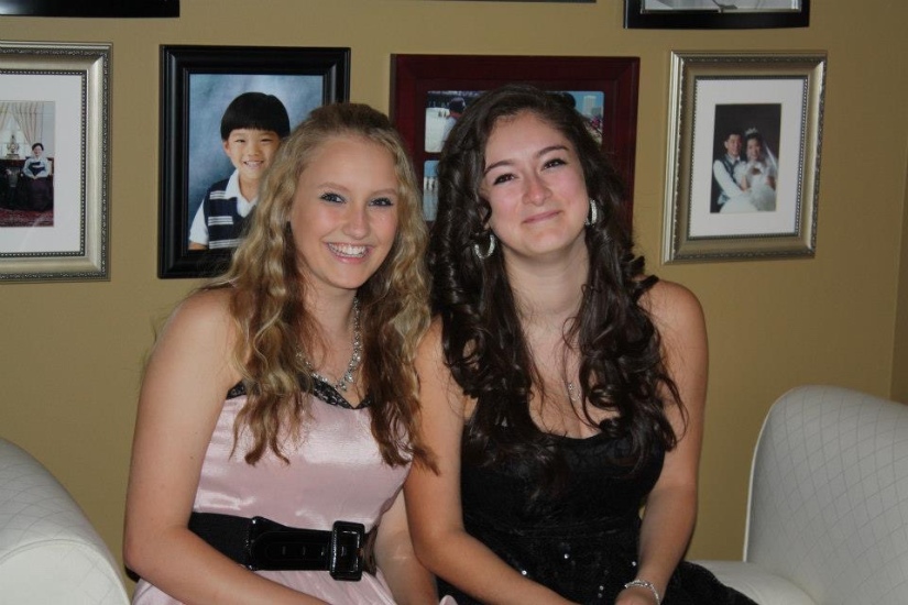 A blonde and brunette girl both dressed up in nice gowns sitting on a couch having homecoming photos taken. 