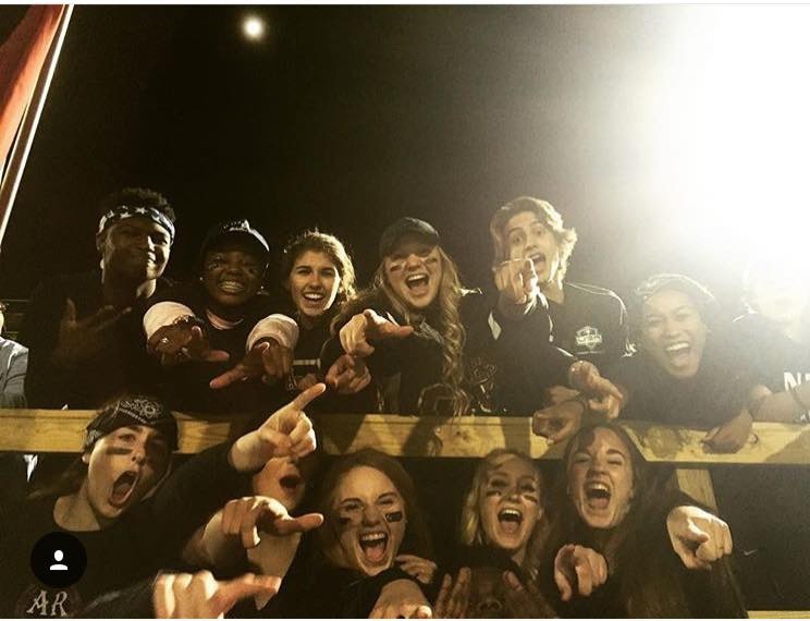 Large group of students all wearing black screaming down into a camera during a football game. 