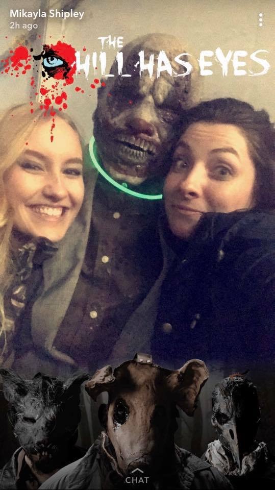 Two girls bundled up in the cold with a person dressed in a mask at a haunted outdoor event.