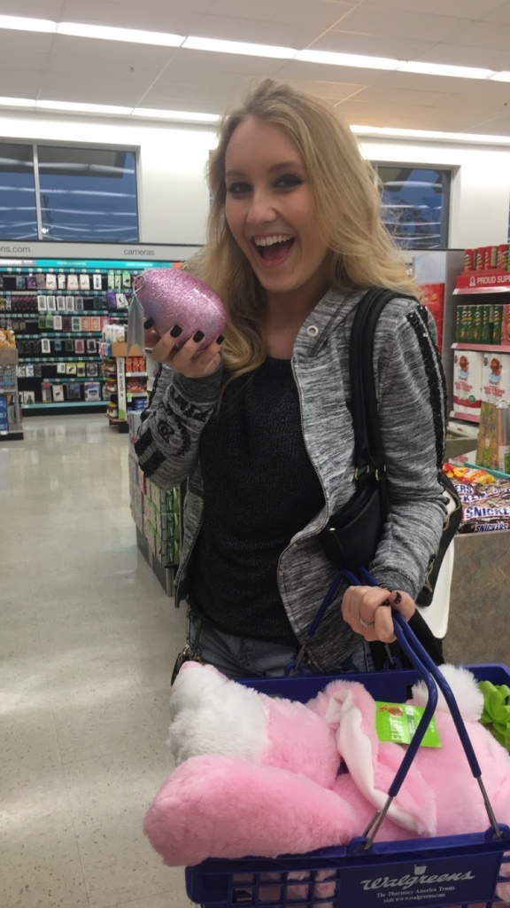 Girl in grey sweatshirt holding a pink glittery Easter egg and a basket with a stuffed pink bunny in it, a very large smile in on her face.  