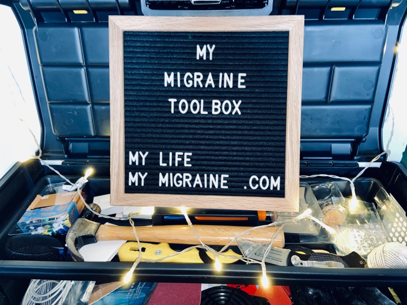 A black felt board with white letters spelling out "my migraine toolbox" propped in an actual toolbox full of hammers and tacks and decorative lights. 