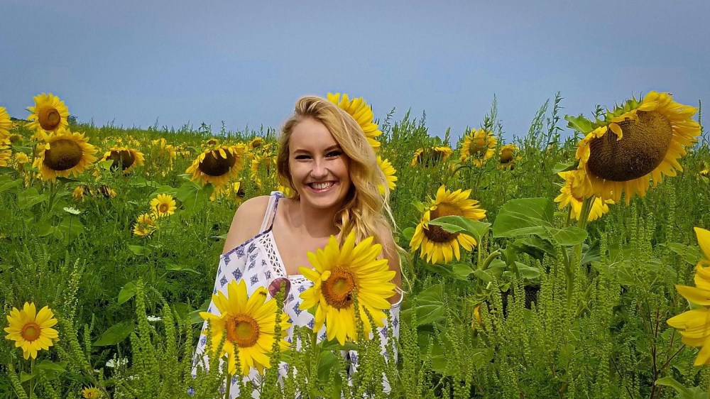 White woman with long blonde hair pulled to one side. She's standing in the middle of a sunflower field wearing a white dress with a small blue pattern. 