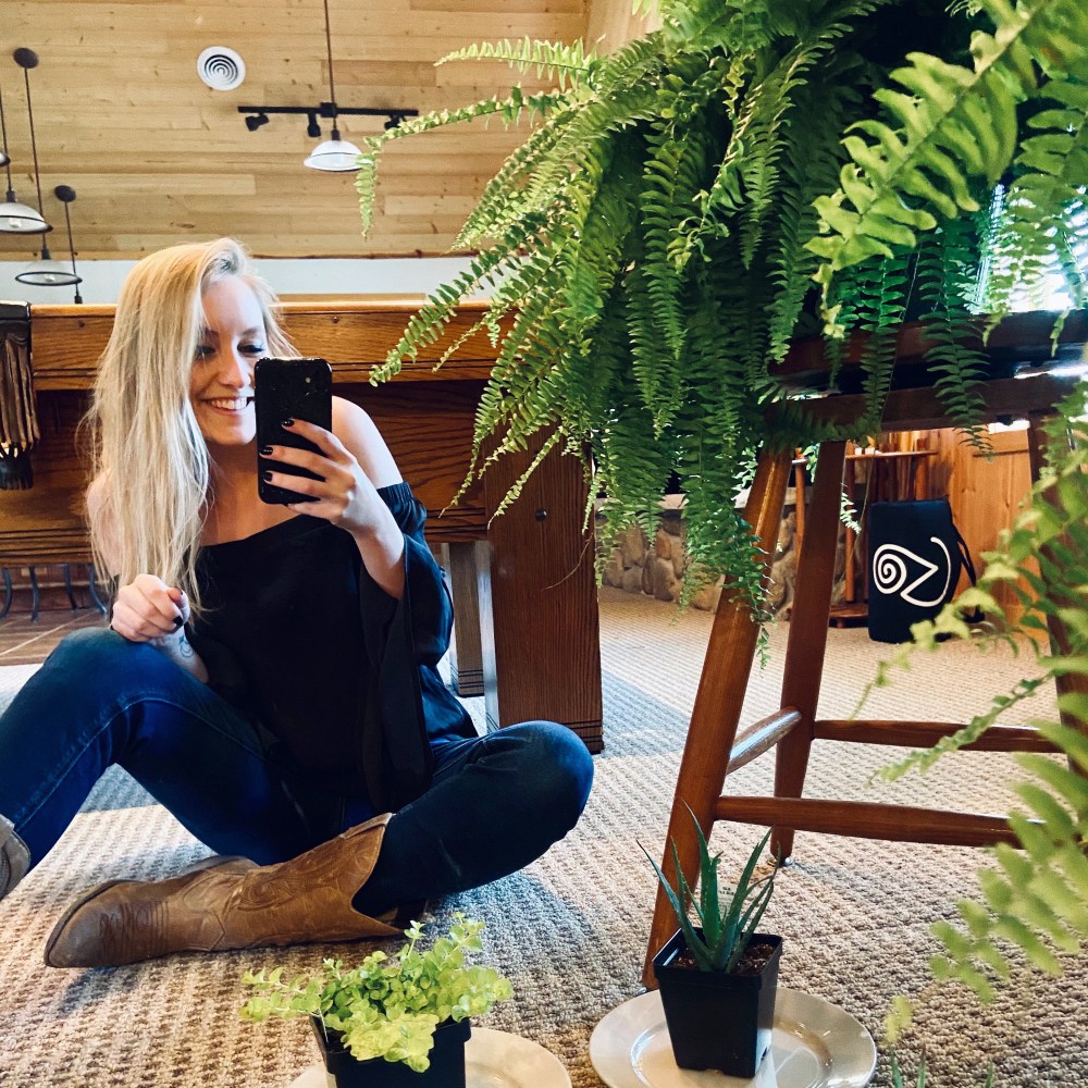 Woman with blonde hair sitting on the floor next to a large fern perched on a stool