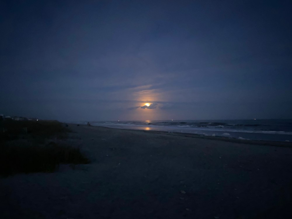 A dimly lit beach with the waves rolling in. A full moon peaks out from behind some clouds. 