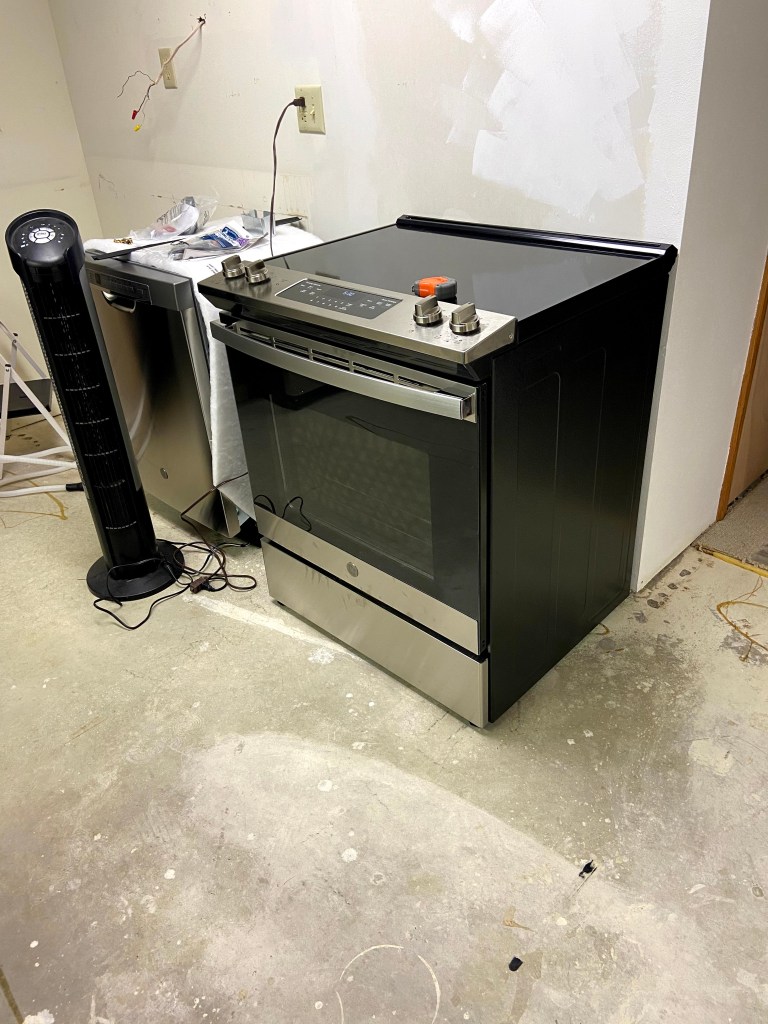 Stainless steel electric stove with front controls installed where door once was, and a new dishwasher sitting next to it. 