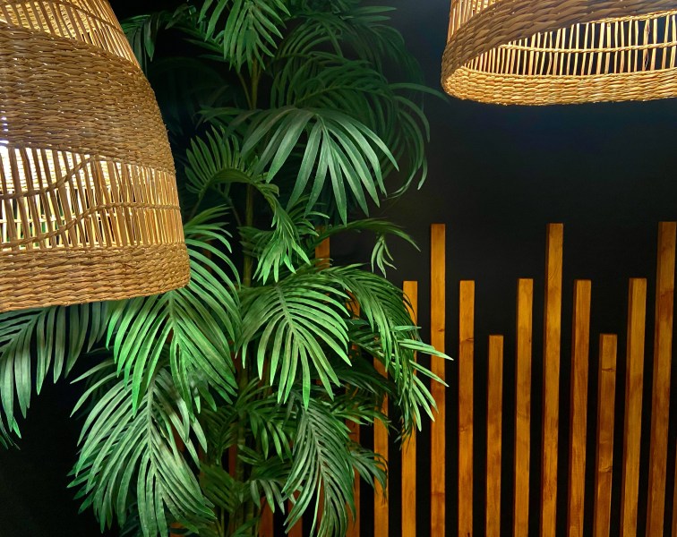A detail shot of the corner of the room, showing the corners of the light rattan pendant shades illuminated against the deep green faux palm. This contrasts with the right side of the photo where the varying height wood boards contrast against the black walls creating an art deco pattern accent wall.