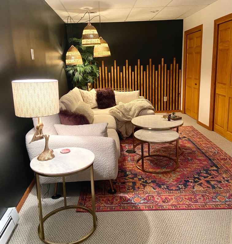 A view of the completed corner of my living room, the far back wall has been completed with the wooden board art deco pattern, a palm tree sits in the corner where three large rattan hanging pendants drape across the ceiling and hang over the corner of the large white boucle sectional. A matching gold and marble round end table has been added to the near side of the couch to match the coffee table. A gold peacock lamp sits atop this end table with a white and gold art deco patterned shade. The space is pulled together with the pink oriental rug and deep eggplant colored Mongolian fur pillows and white pillows adorning the couch.