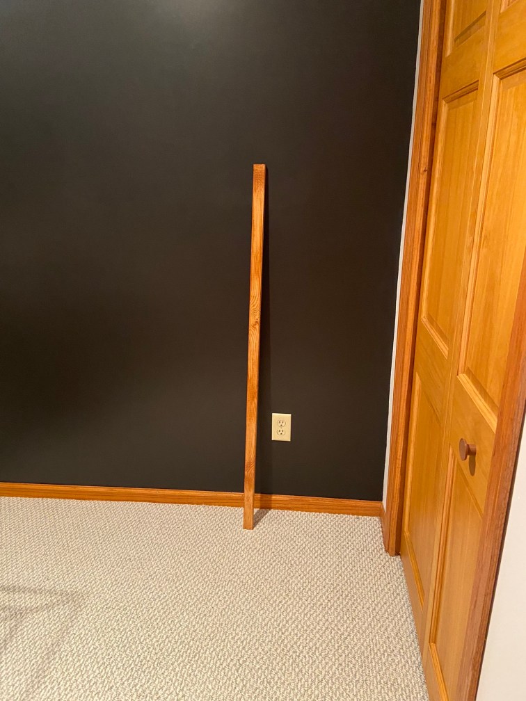 A single piece of 1 by 2 board that has been cut and stained is propped against the freshly painted black walls. The stain of the board matches the wood trimmed doors to the right and the wood trimmed baseboard perfectly. It's a warmer wood tone, perhaps slightly orangish in color which pops dramatically against the black paint.