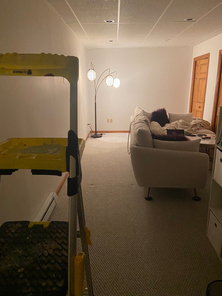 My living room pre paint set up. My white sectional has been pulled to the right side of the room leaving the far wall and the long wall to the left - both white - completely accessible. A ladder stands at the front of the frame.