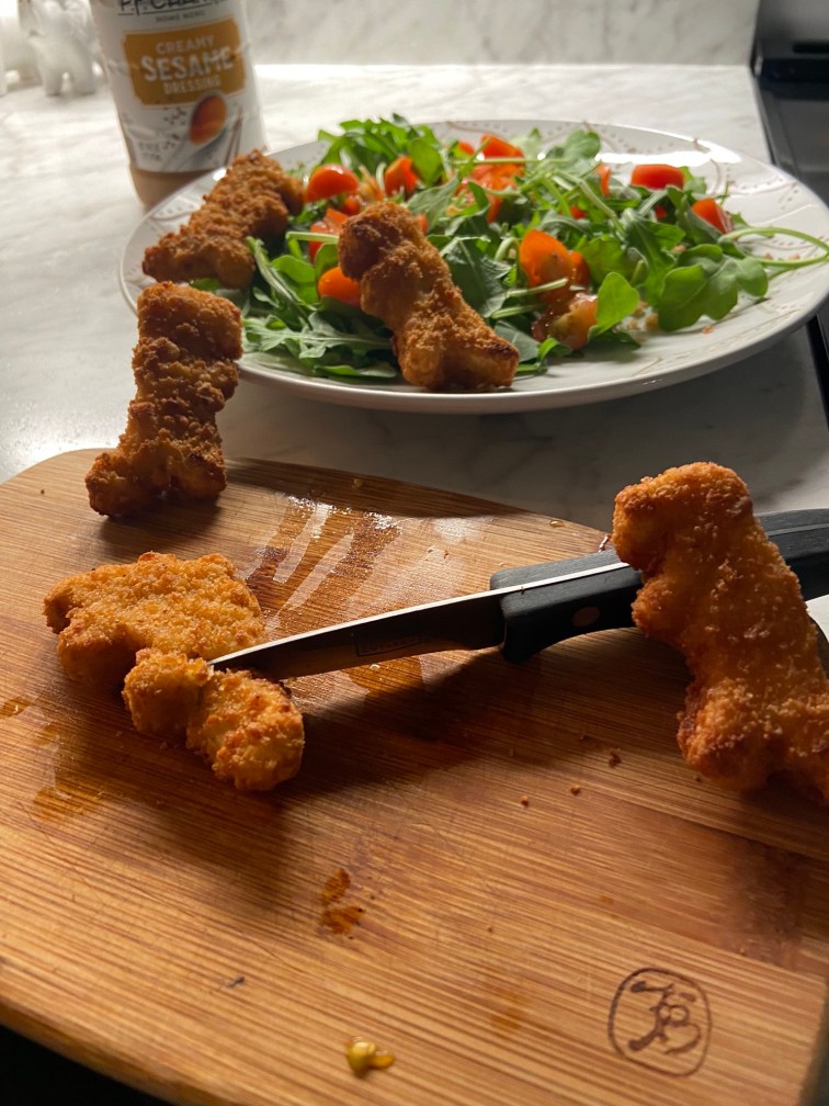 Dinosaur chicken nuggets hold a small knife in their hand as the chop up other dinosaur chicken nuggets on a cutting board. Some of the nuggets attempt to escape to the arugula and tomato salad behind it.