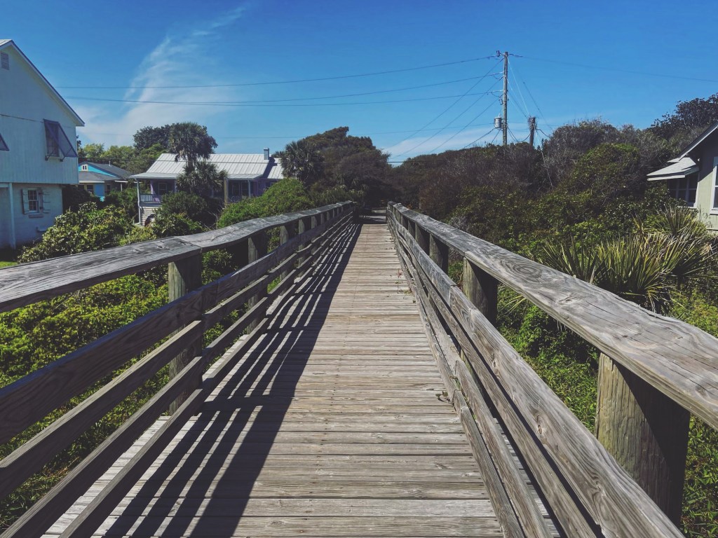 A long path leading away from the beach. The path is a wooden bridge that extends out as far as the eye can see, tropical foliage lining the sides of each bath with small beach cottages peaking out from behind palm trees along each side of the bridge. The sky above is blue. The image exudes a sense of calm.