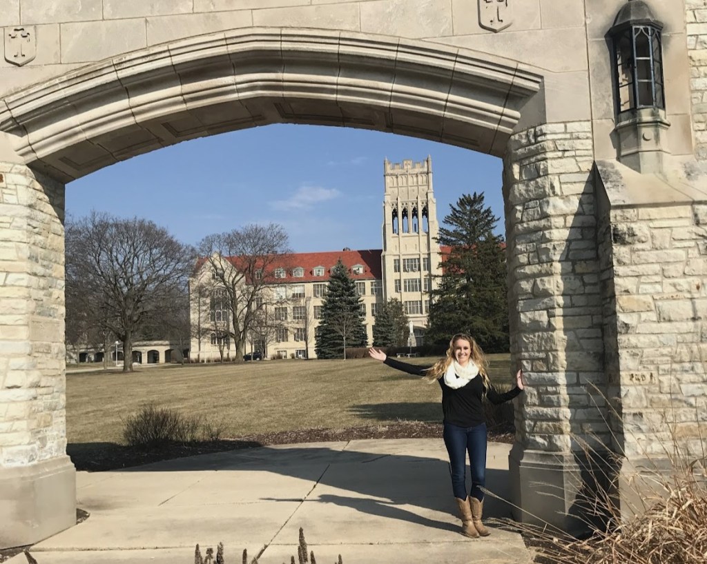 A photo of me, a blonde white woman, standing in front of a small catholic university my arms extended upward in celebration.