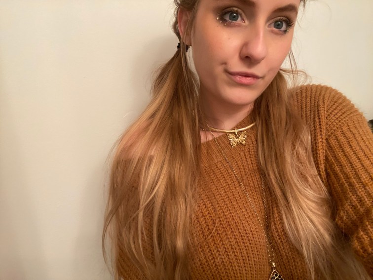 Me. A blonde white woman. I’m wearing a caramel colored sweater with a gold butterfly necklace. My hair is braided in French braids that let loose into two pig tails. I have a smoky eyed makeup look on completed with some gemstones.