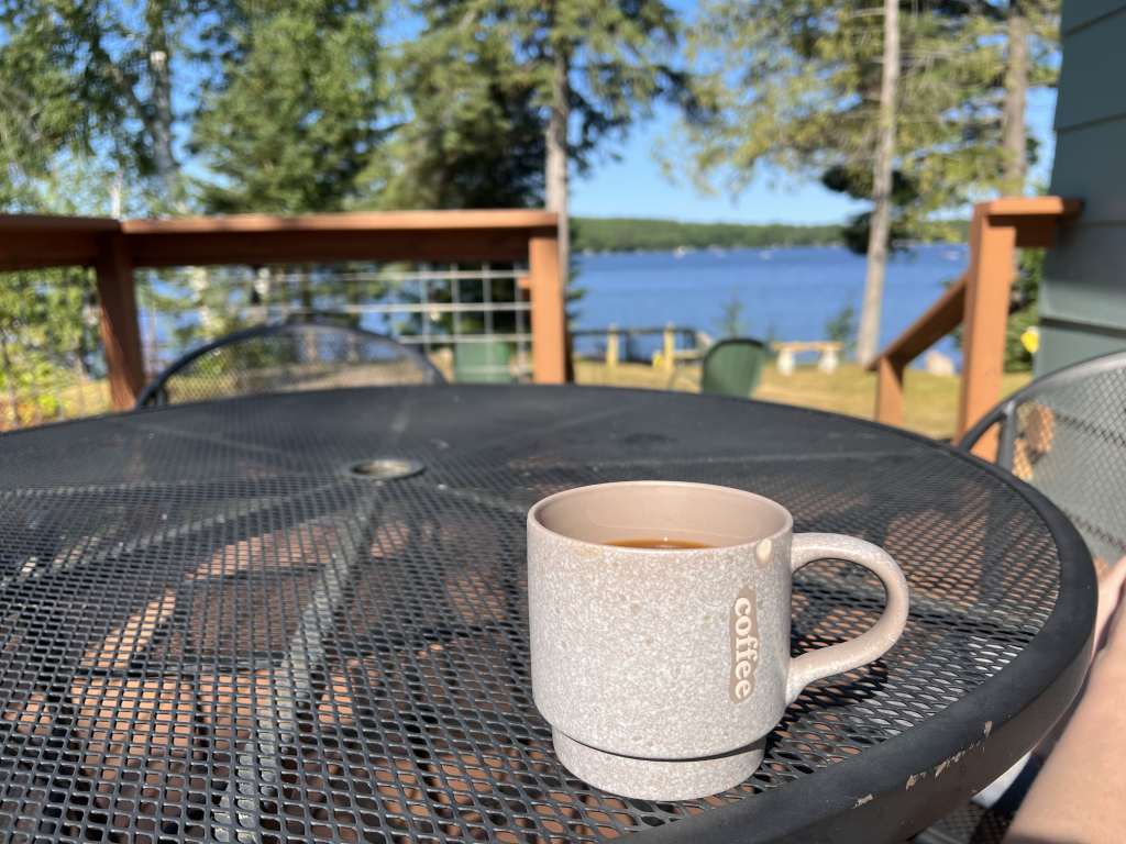 An in focus beige coffee mug sitting on a black patio table with the sun shining and the lake shimmering in the distance.