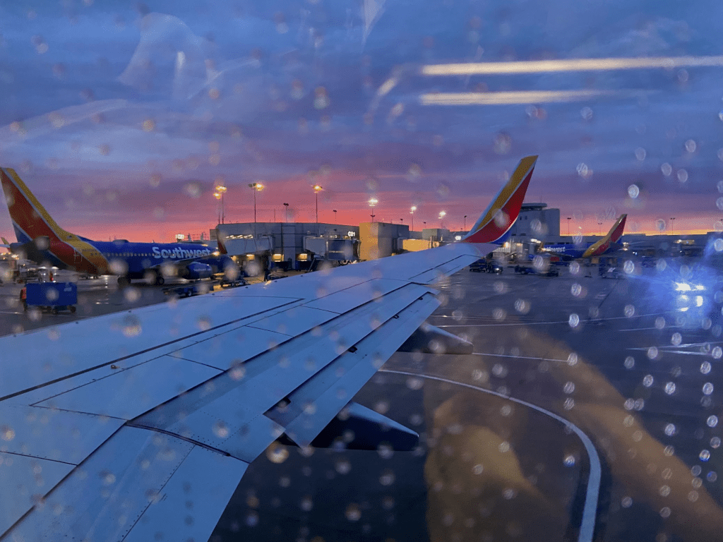 The view out my airplane window, streaks of rain blocking some of the view. I'm sat on the wing, southwest planes docked across from the window with a stormy sky above fading to vibrant pink on the horizon.