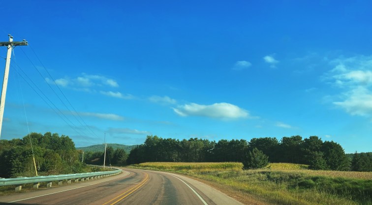 A winding road stretches across the mountainous rural countryside curving to the left. The sky is blue