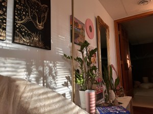 A soft sunlit scene of my coffee lounge. The sunlight hits the wall against where I’m seated in my papasan chair, casting shadows across golden artwork and stacks of books and plants.