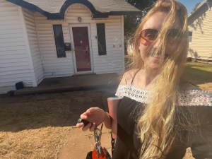 a selfie i took of me, a blonde white woman wearing sunglasses and a black and white shirt smiling in front of a small white house with a red door and a circular arch in the roof over the doorway